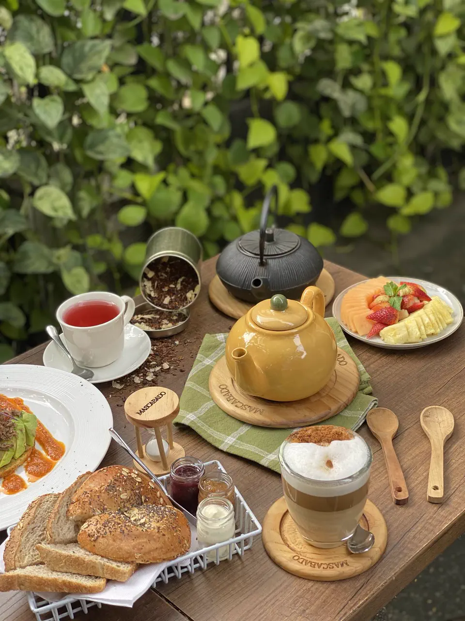Mesa de té en la terraza con la tetera amarilla, pan de la casa, fruta y un capuchino entre plantas
